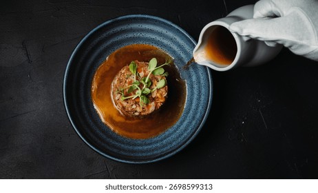 White-gloved hand pouring rich brown sauce onto a garnished dish in a dark blue plate